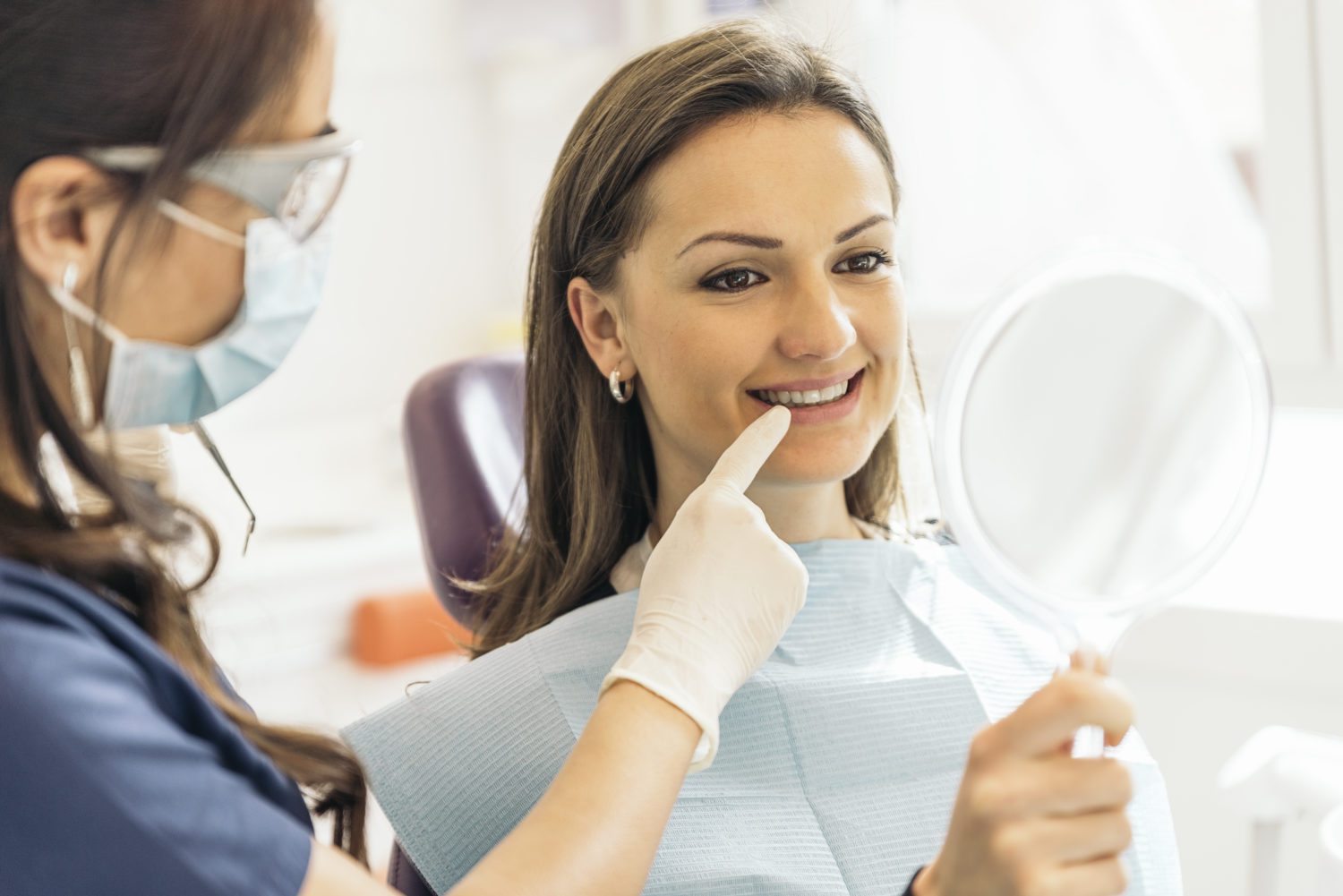 Young woman looking into a handheld mirror as a dental hygienist points at her mouth